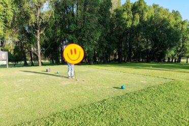 A golfer standing on a lush, well-maintained turf grass tee box, preparing to hit a golf ball, with a dense line of trees in the background.