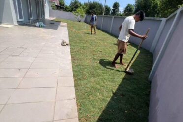 Two workers laying and tending to new turf grass in a long, narrow side yard between a modern grey house and a tall white perimeter wall on a sunny day.