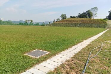 A large recreational field with newly installed turf grass, featuring a long concrete drainage path and a utility grate in the foreground, with a grassy slope and urban skyline in the distance.