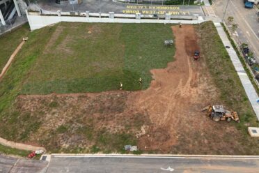 Aerial view of a construction site showing workers and heavy machinery laying down turf grass (sod) on a large, mostly bare dirt field next to a paved road and building.