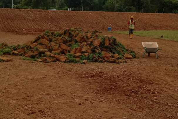 A large pile of turf grass (sod) rolls dumped on a vast, flat expanse of reddish-brown construction dirt, with a worker and a wheelbarrow nearby.
