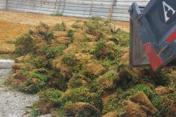 A pile of turf grass sod rolls being dumped from the back of a black dump truck onto a gravel and dirt staging area next to an exposed metal sheet barrier and construction equipment.